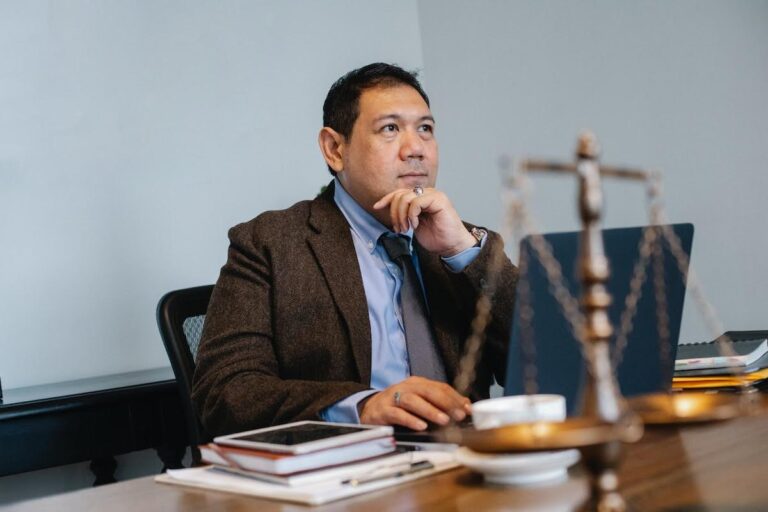Business litigation lawyer sitting at a desk, thoughtfully reviewing case details with legal scales, a laptop, and documents in a professional office setting.