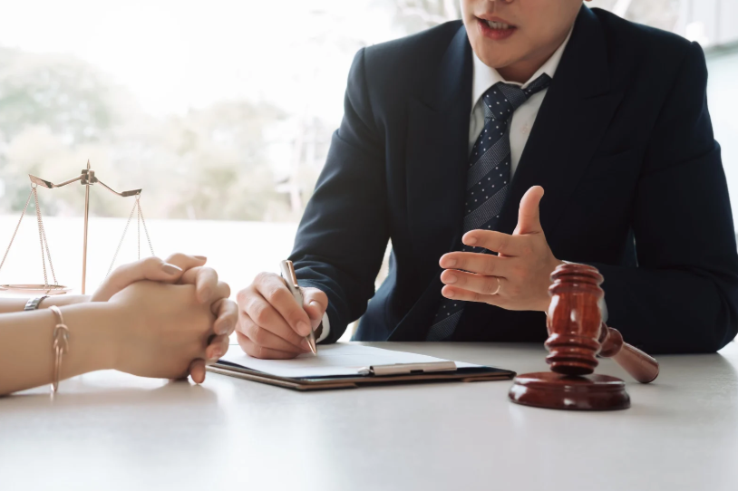 A lawyer discusses terms with a client while a gavel and scales of justice are present on the table. This scene indicates legal discussions and advice about a non-compete agreement, advising the client on the restrictions and enforceability of the agreement.