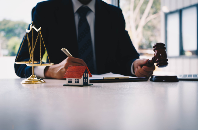 A gavel, scales of justice, and a model house on a desk represent the legal aspect of title insurance. This imagery emphasizes that title insurance offers protection during property transactions, ensuring the validity of ownership and protecting against unforeseen legal issues after the purchase.