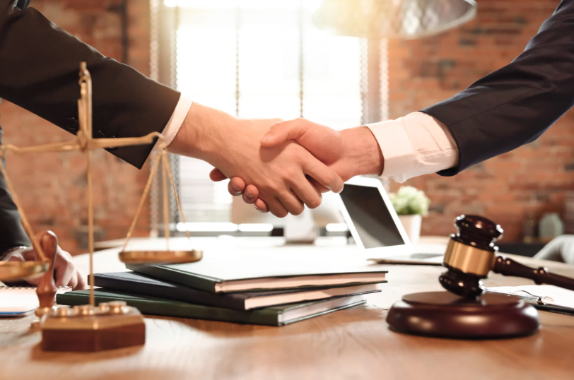 Two professionals shaking hands near legal documents, scales of justice, and a gavel, representing the services of a business attorney.