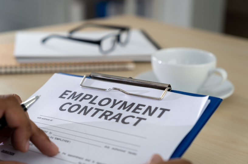 Employment contract document on a clipboard being reviewed, with office items like glasses, a notebook, and a coffee cup in the background, symbolizing the services of an employment contract lawyer.